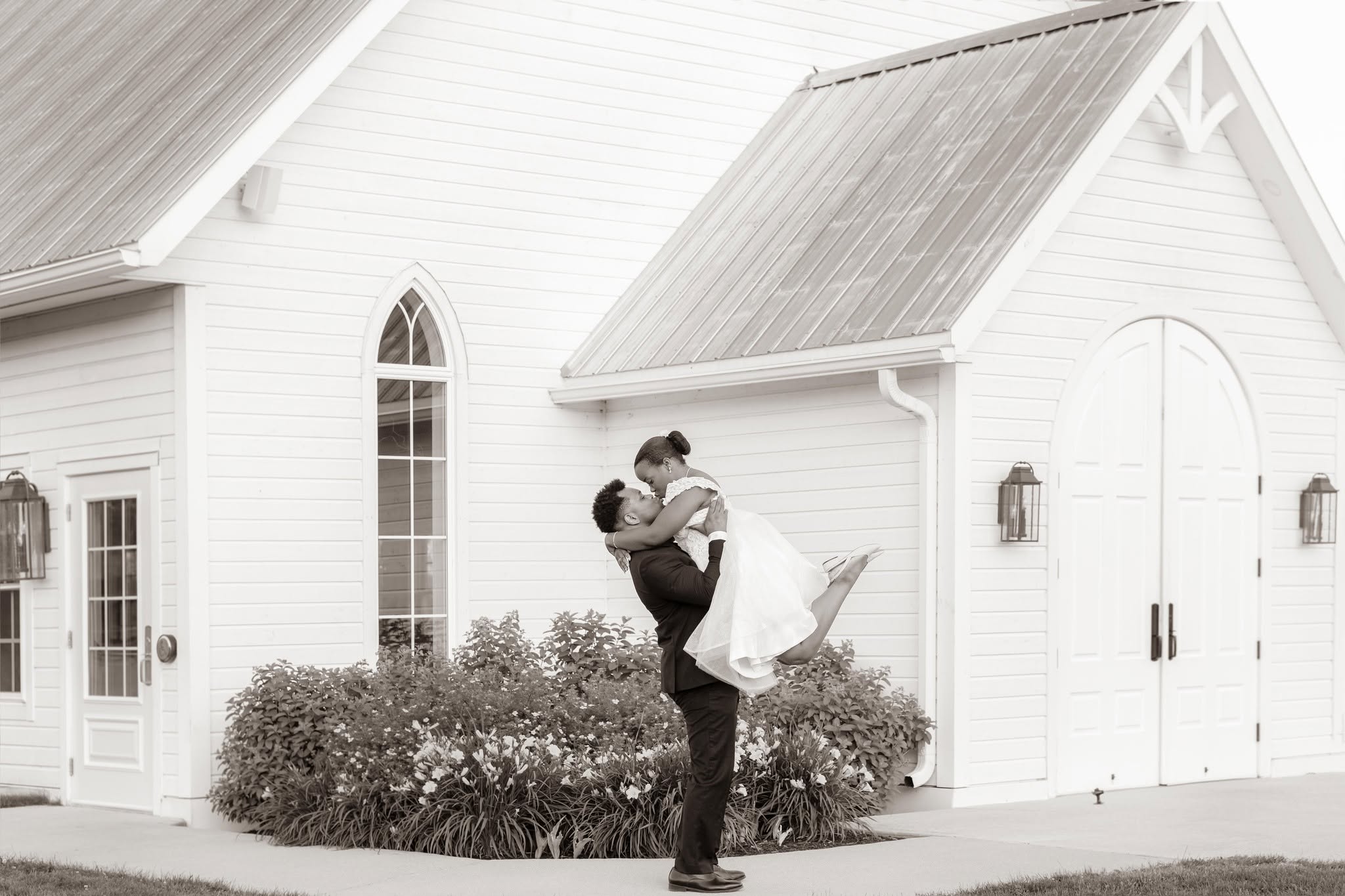Un marié soulève sa mariée devant une chapelle blanche — photographie de Reginald David